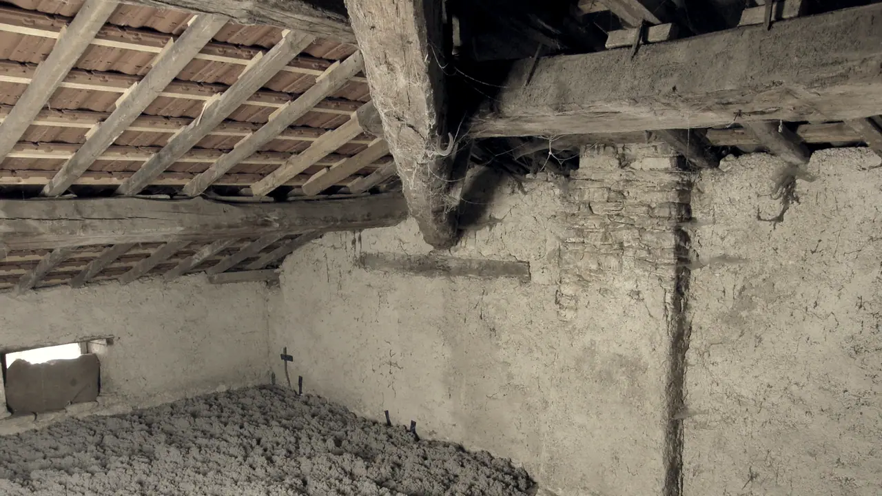 Grenier d'une maison ancienne sous sa charpente de bois et ses tuiles, avec au pied des murs enduits une épaisse couche d'isolant soufflé sur le plancher.