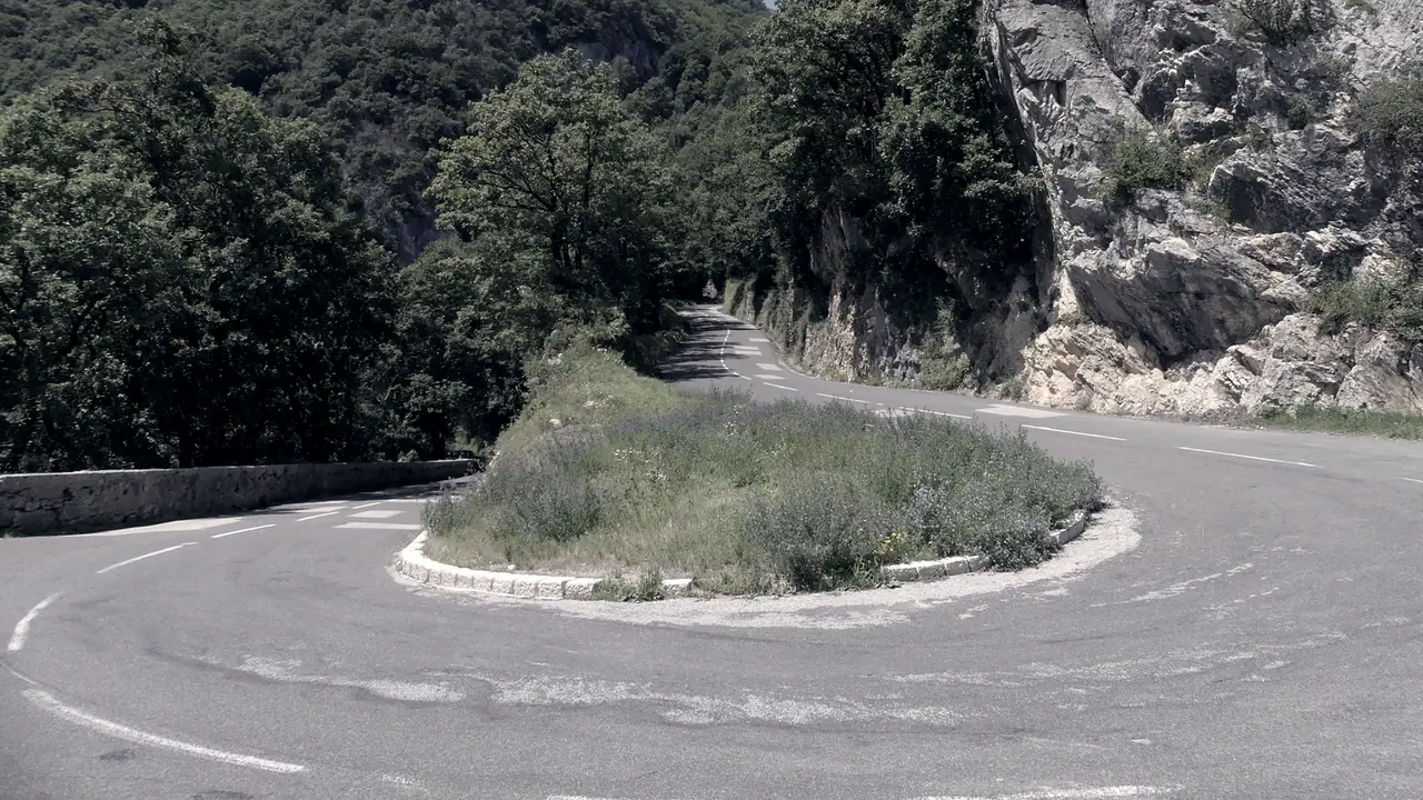 Virage en épingle d'une route de montagne qui repart vers le haut, entre une paroi de calcaire et des pentes boisées, autour d'un îlot d'herbes folles.