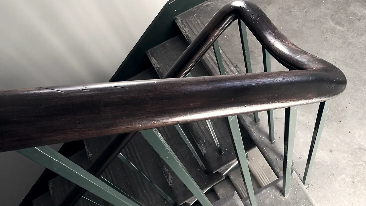 Main courante de bois sombre, polie par l'usage, qui tourne au-dessus d'une cage d'escalier ancienne aux barreaux peints en vert.