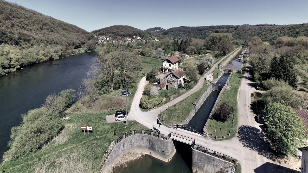 Vue plongeante sur une rivière et un canal qui courent côte à côte dans une vallée boisée, avec au premier plan les murs d'une écluse enjambée par une passerelle.