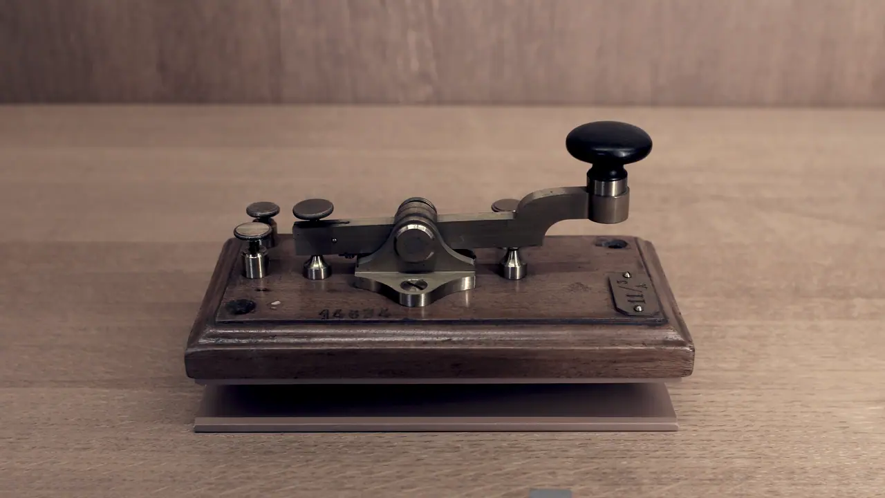 Manipulateur de télégraphe Morse ancien exposé sur une étagère de musée : un levier de laiton à pommeau posé sur un socle de bois mouluré, avec ses bornes de raccordement.
