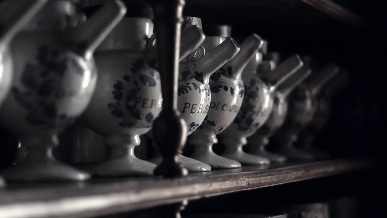 Rangée de pots à pharmacie anciens en faïence blanche peinte de feuillages, posés sur une étagère de bois sombre, le bec tourné vers le côté.
