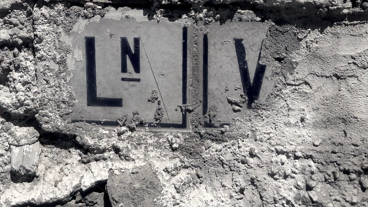 Deux petites plaques émaillées scellées dans un crépi, marquées LN et V en grandes lettres sombres, de part et d'autre de la limite entre deux communes.