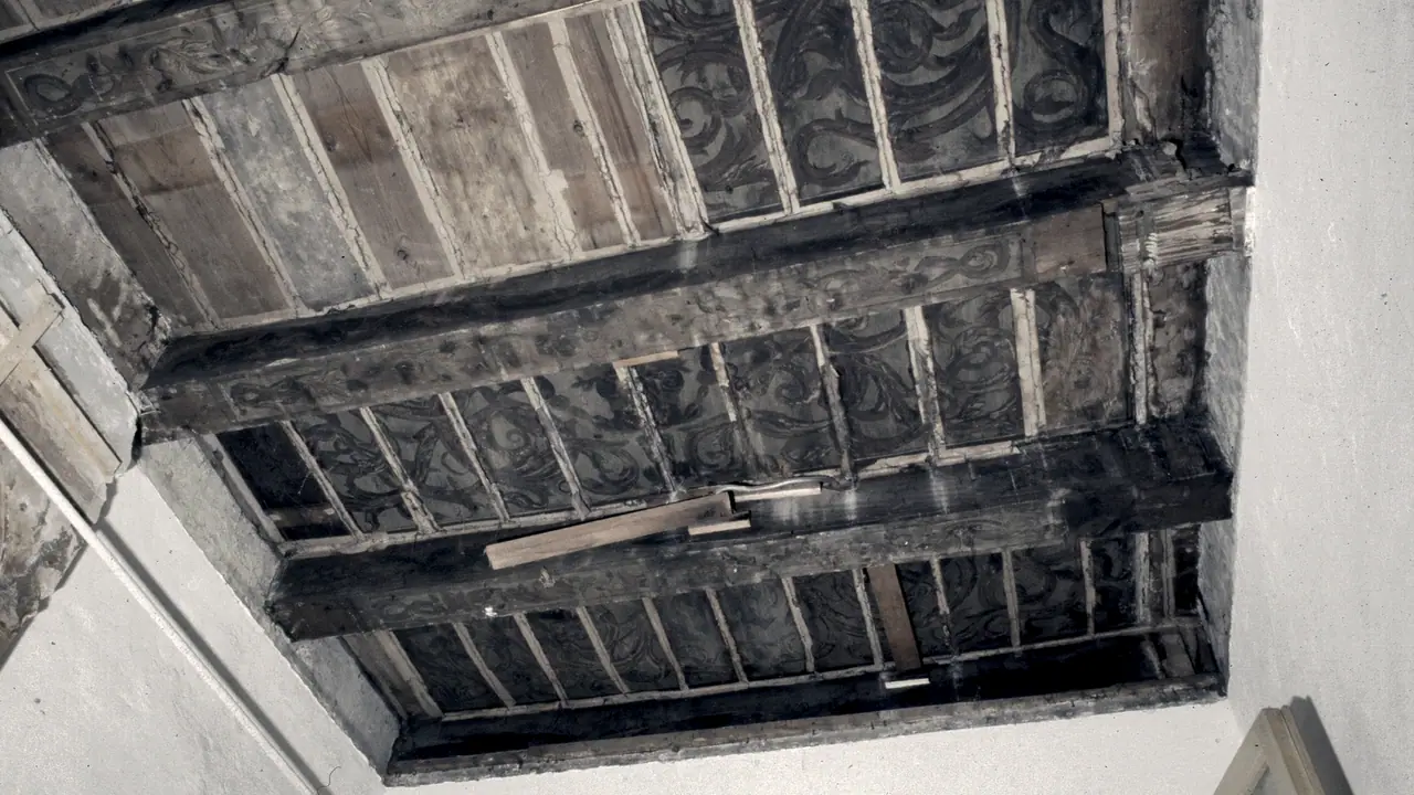 Plafond ancien à poutres de bois vu d'en bas, ses entrevous peints de rinceaux sombres, entre deux murs blanchis.