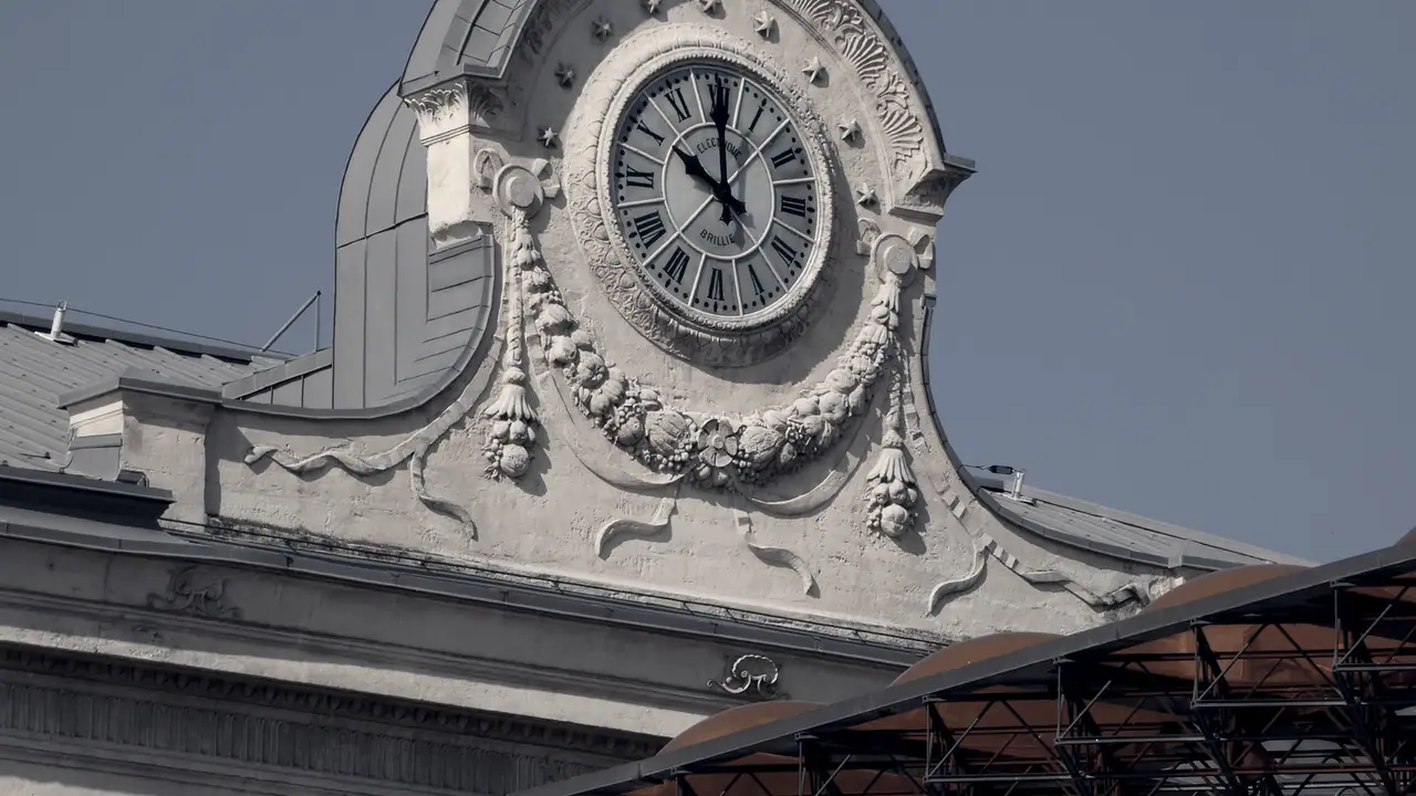 Grande horloge à chiffres romains encastrée dans le fronton sculpté d'un bâtiment, au-dessus d'une toiture métallique.