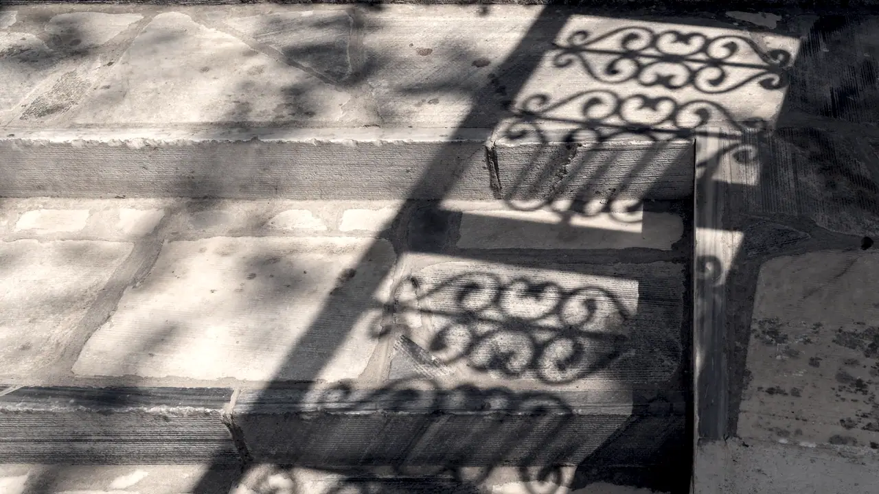 Ombre d'une grille en fer forgé projetée sur des dalles de pierre claire, motifs de volutes découpés par la lumière.