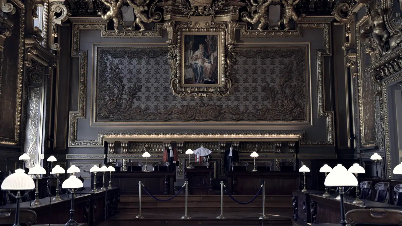 Salle d'audience de la Cour de cassation aux boiseries sculptées et dorées, avec ses lampes alignées sur les bancs et des robes de magistrat exposées derrière le siège des juges.