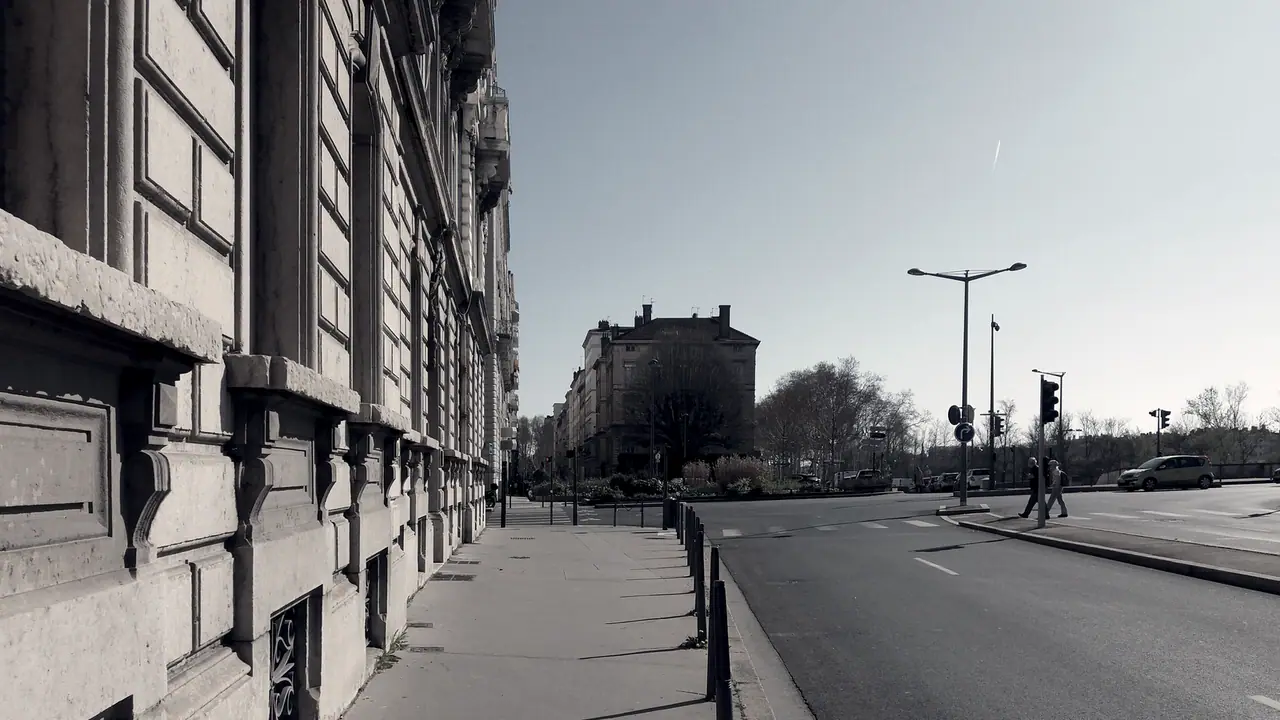 Trottoir longeant la façade de pierre d'un immeuble ancien aux consoles sculptées, avec au bout du quai un autre immeuble à l'angle d'un carrefour.
