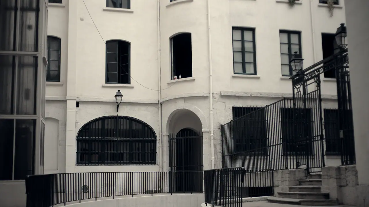 Cour intérieure d'un immeuble ancien, avec une grille en fer forgé, une lanterne murale et une volée de marches en pierre sur la droite.