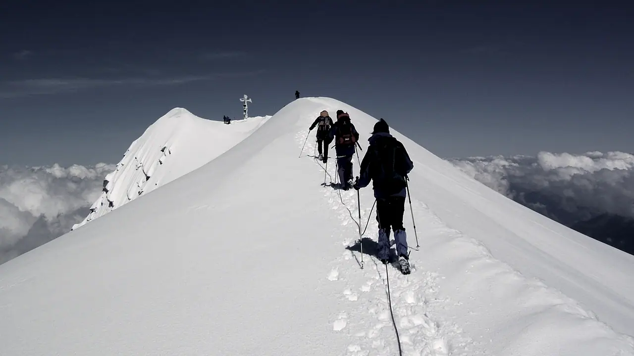 Trois alpinistes encordés montent à la file vers une croix sommitale sur une arête enneigée, vus de dos, la corde courant dans la neige jusqu'au premier plan.