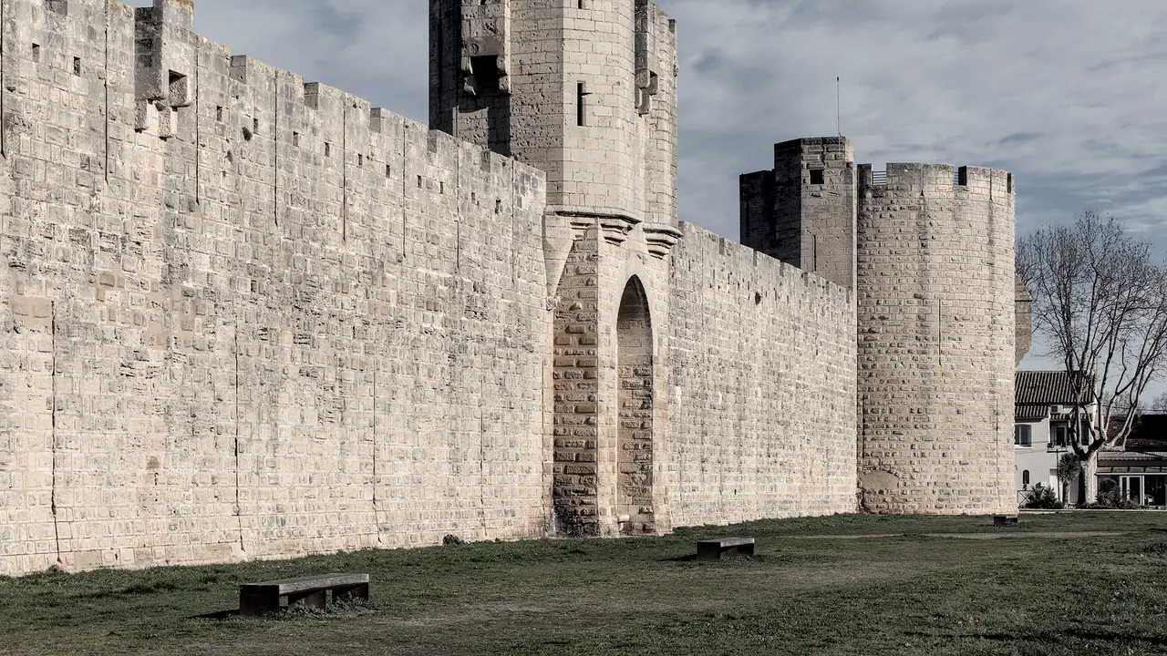 Longue muraille de pierre des remparts d'Aigues-Mortes, percée d'une porte en arc brisé au pied d'une tour carrée, avec une tour ronde plus loin et deux bancs sur la pelouse.