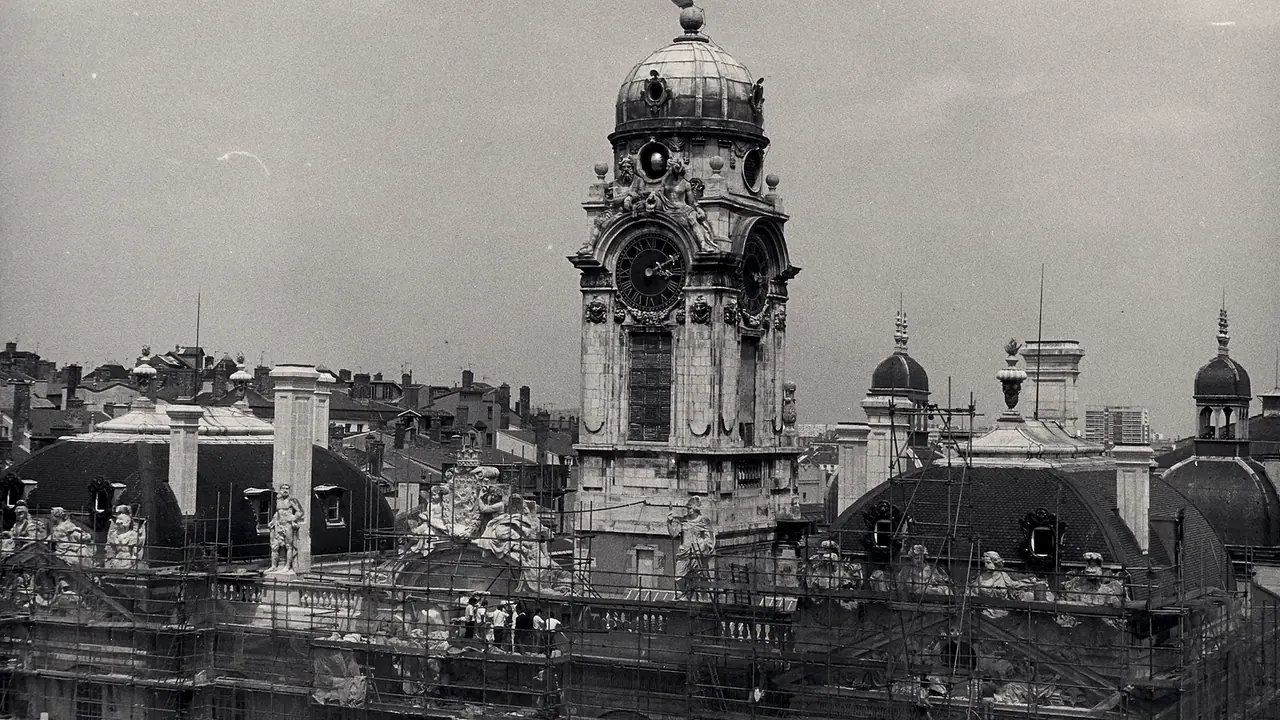 Beffroi d'horloge en pierre sculptée entouré d'échafaudages de tubes, avec des statues et des dômes sur les toits au premier plan.