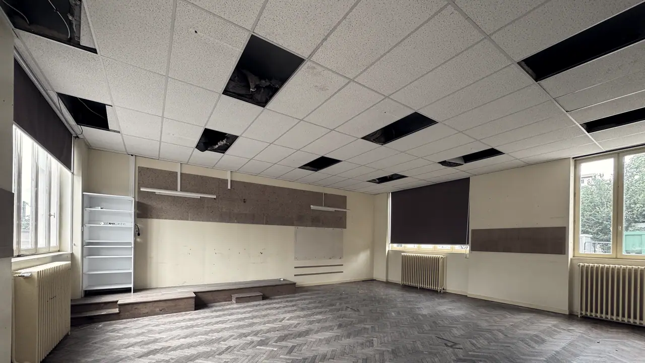 Grande pièce vide avant travaux : dalles de faux plafond déposées, parquet à bâtons rompus usé et radiateurs en fonte sous les fenêtres.
