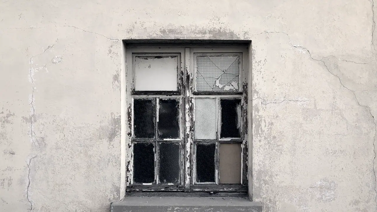 Fenêtre ancienne aux carreaux cassés et au bois écaillé, dans un mur d'enduit clair fatigué par les intempéries.