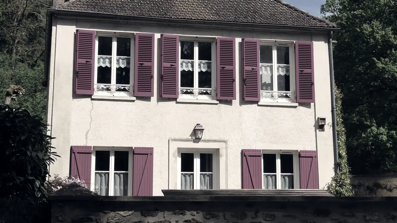 Façade de maison de village avec ses six fenêtres à volets ouverts, ses rideaux de dentelle, sa lanterne et le mur de clôture devant elle.