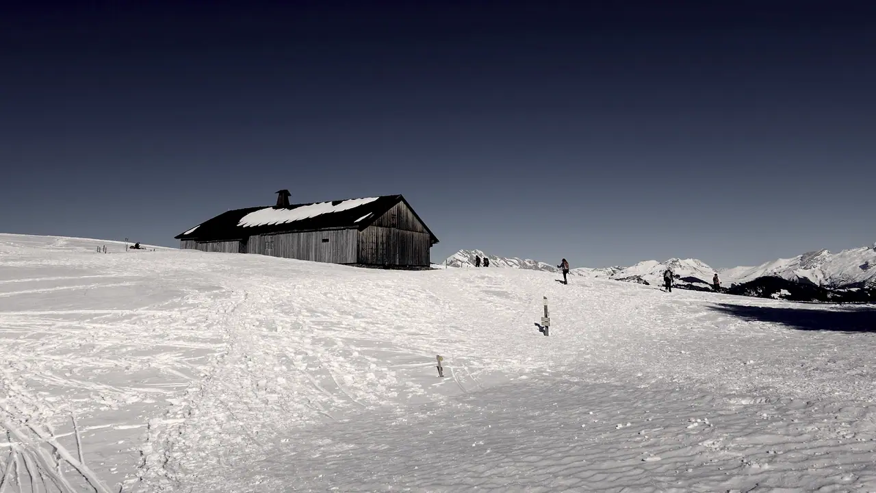 Chalet de bois isolé au milieu d'un alpage couvert de neige, avec ses traces de ski et la ligne des sommets derrière lui.