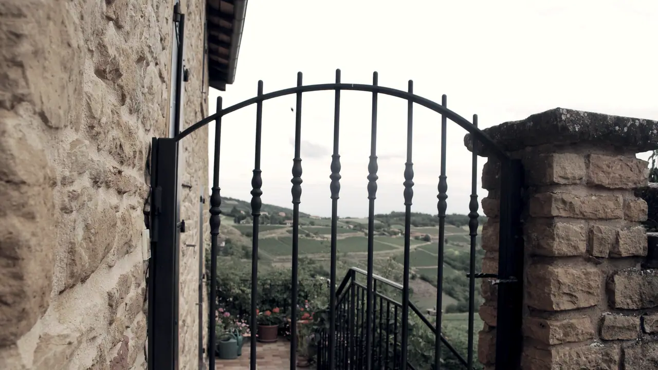 Grille de fer ouvragée entre deux murs de pierre, ouverte sur une terrasse et sur des coteaux plantés de vignes jusqu'à l'horizon.