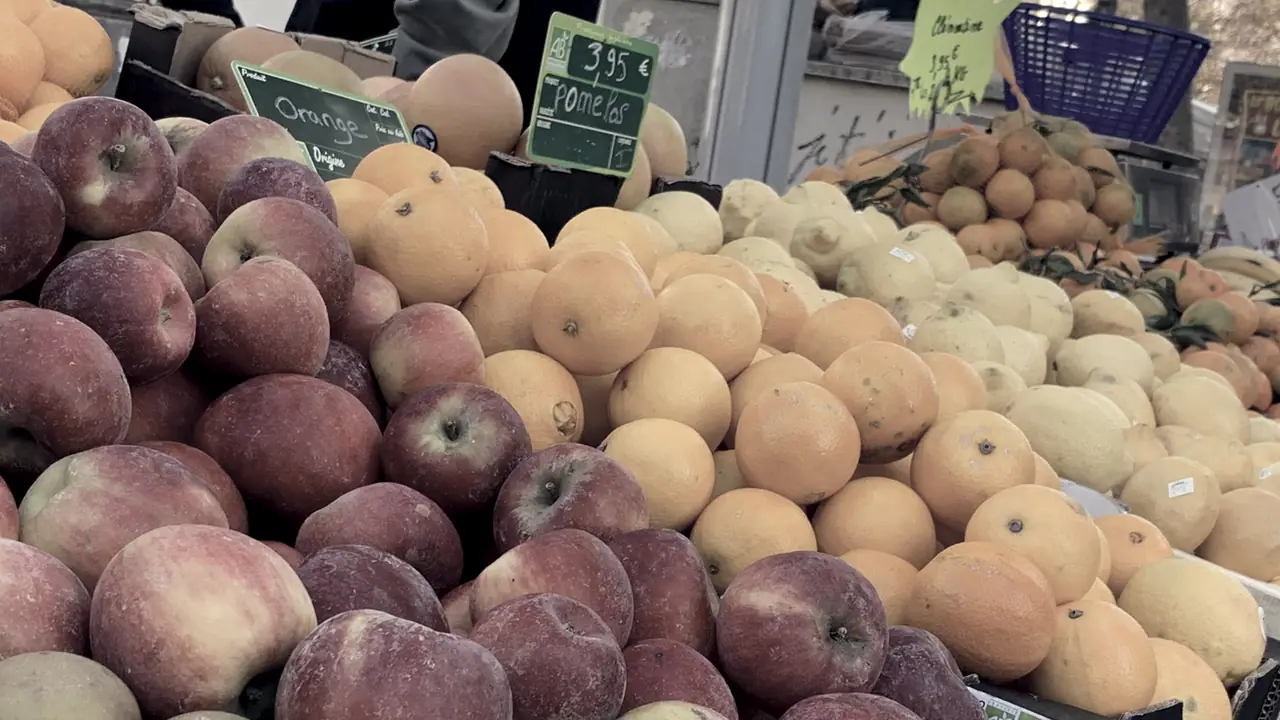 Étal de marché couvert de pommes et d'agrumes empilés dans des cageots, avec des ardoises de prix manuscrites au-dessus des fruits.