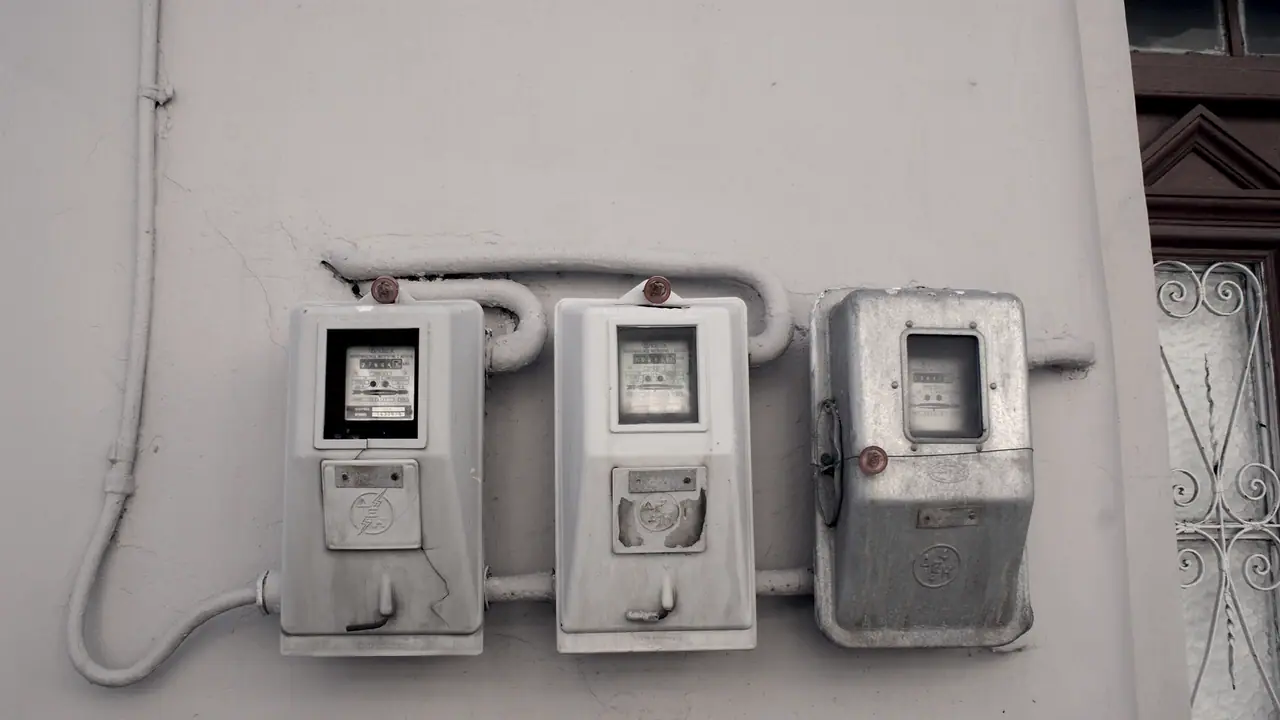 Trois compteurs électriques fixés côte à côte sur un mur crépi, avec leurs gaines apparentes et une grille de fenêtre ouvragée.