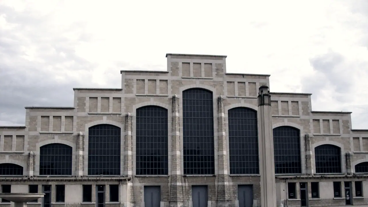 Façade symétrique d'une halle industrielle en pierre, avec de hautes verrières cintrées et un lampadaire dressé devant, sous un ciel nuageux.