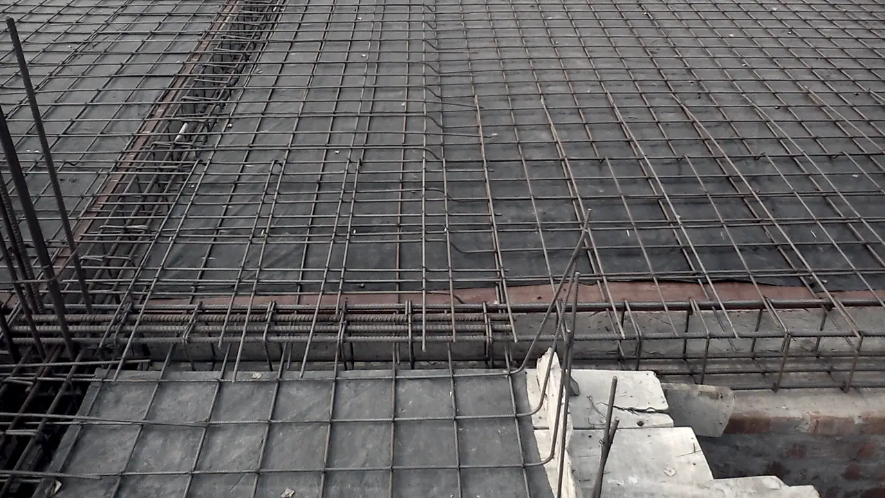 Treillis d'armatures métalliques posé sur le coffrage d'une dalle en construction, avec des fers en attente dressés sur le bord et des blocs de béton au premier plan.