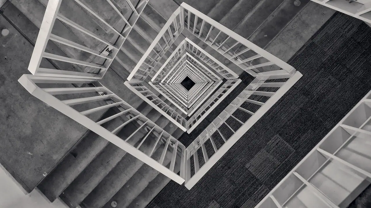 Cage d'escalier carrée vue en plongée, avec des rampes claires qui s'emboîtent en spirale jusqu'au petit carré sombre du fond.