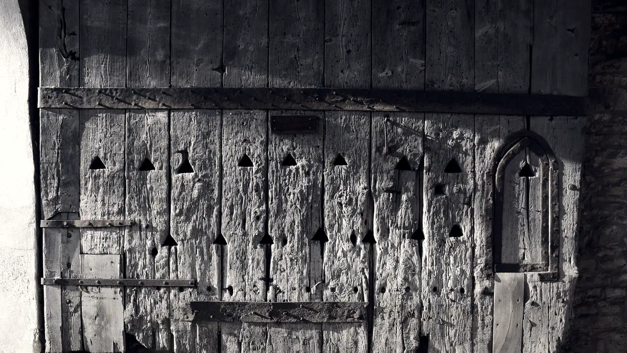 Vantail d'une porte ancienne en bois gris délavé, percé d'ouvertures triangulaires, avec une petite porte en arc brisé découpée dans le battant.
