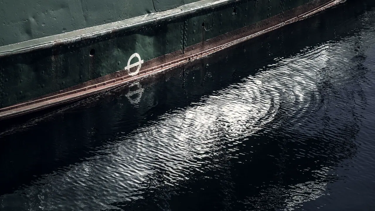 Flanc riveté d'une coque de navire, avec son disque de franc-bord peint juste au-dessus de la ligne de flottaison et son reflet brisé dans l'eau du port.