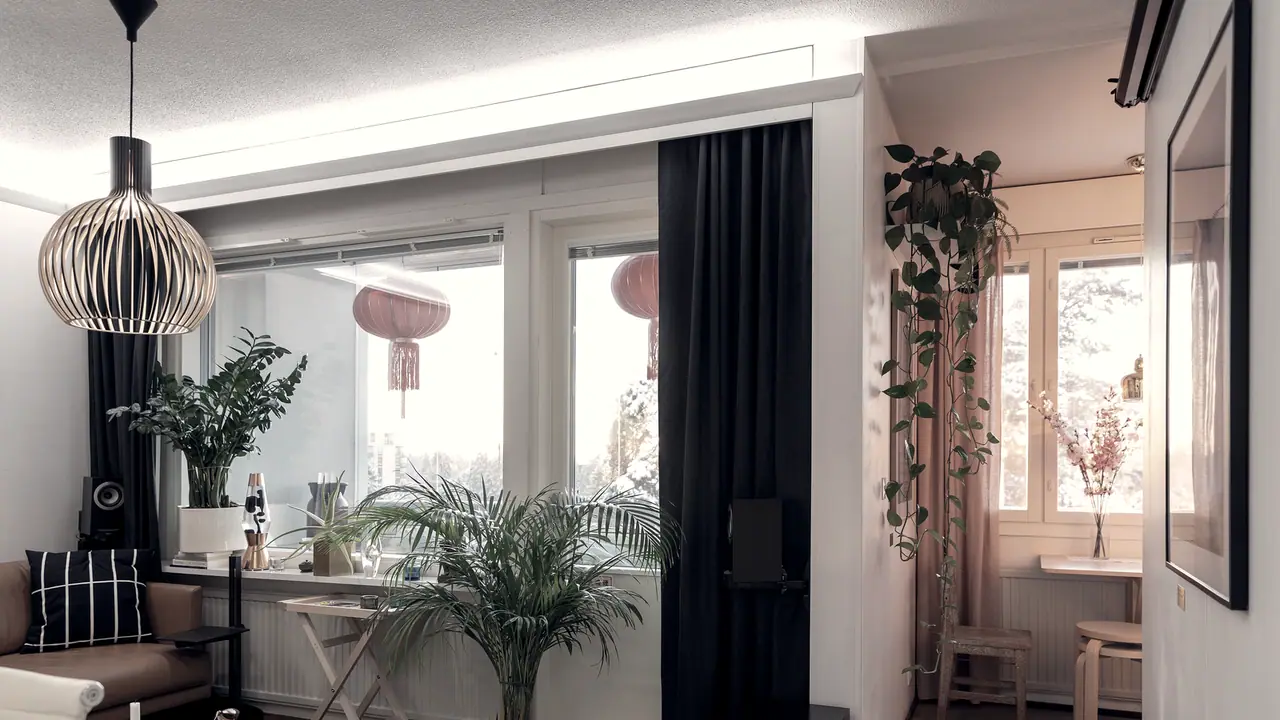 Pièce de séjour meublée avec sa suspension, ses plantes en pot, son fauteuil et ses grandes fenêtres habillées de rideaux.