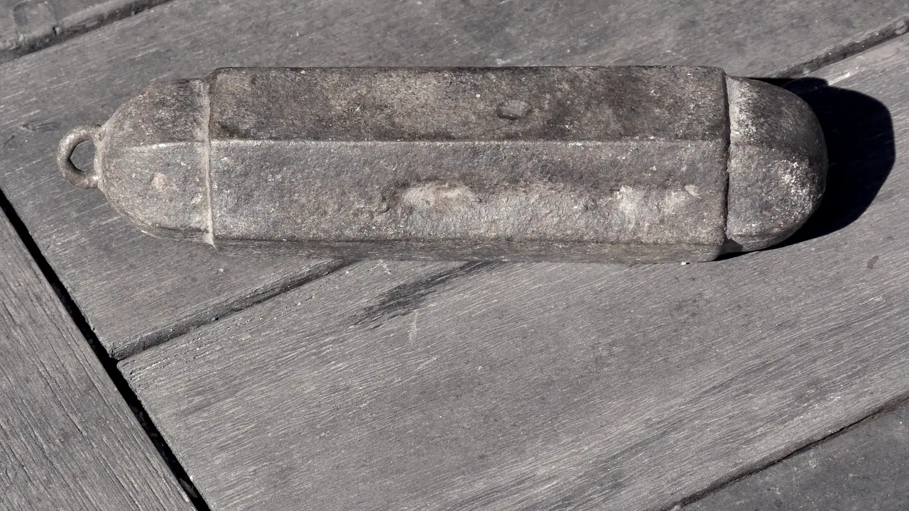 Poids d'horloge en fonte piquée, avec son anneau de suspension, posé à plat sur des planches de bois grisées par le temps.