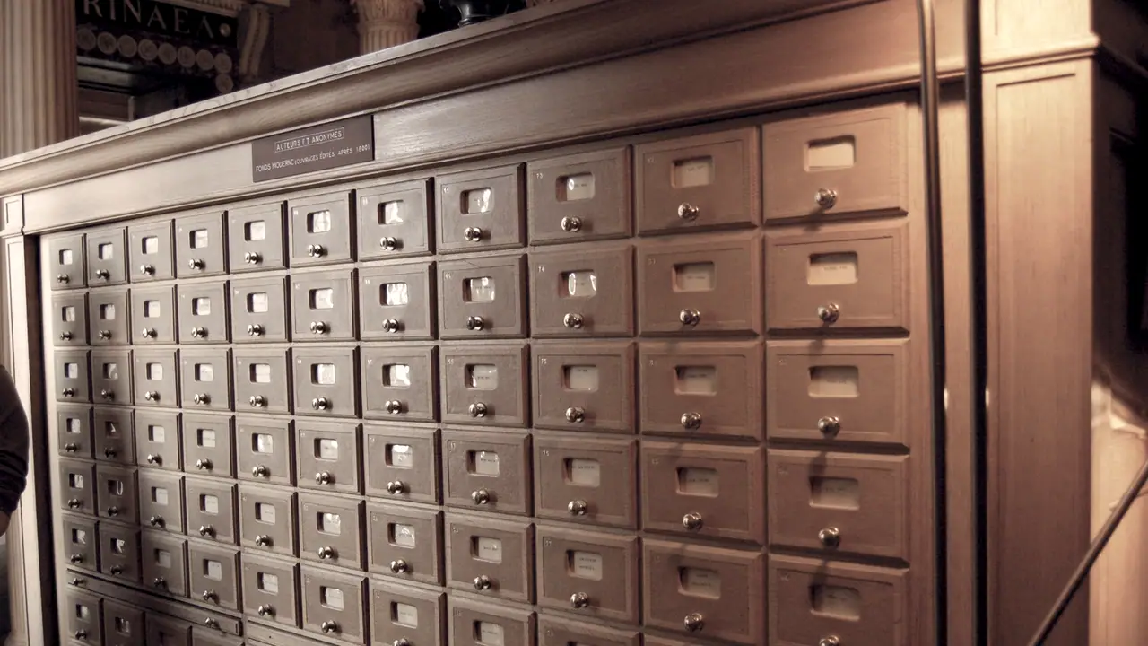 Meuble à fiches de bibliothèque : des dizaines de petits tiroirs de bois alignés, chacun avec son étiquette sous un cadre de laiton.