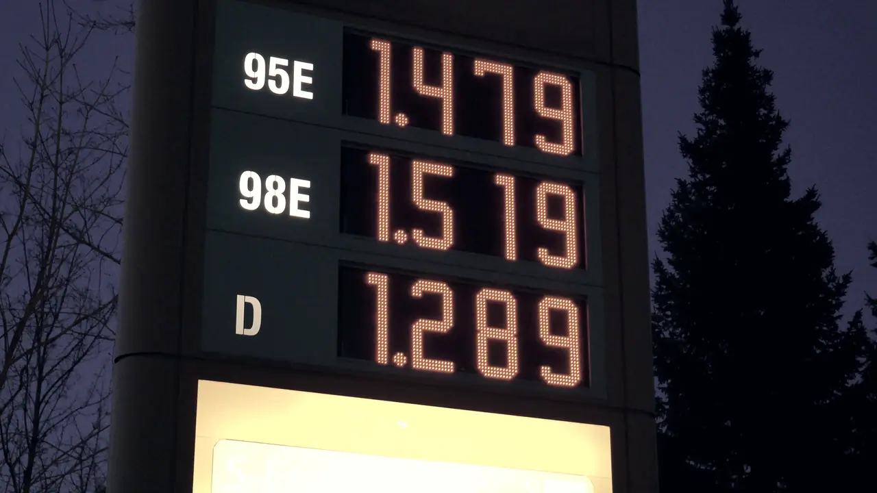 Totem de prix de carburant à la tombée de la nuit : trois lignes de chiffres lumineux, chacune en face d'un code de produit, devant la silhouette d'un sapin.