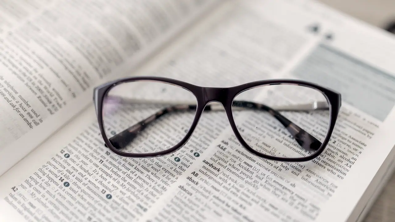 Paire de lunettes posée sur un dictionnaire ouvert, les branches repliées au-dessus de colonnes de définitions en petits caractères.