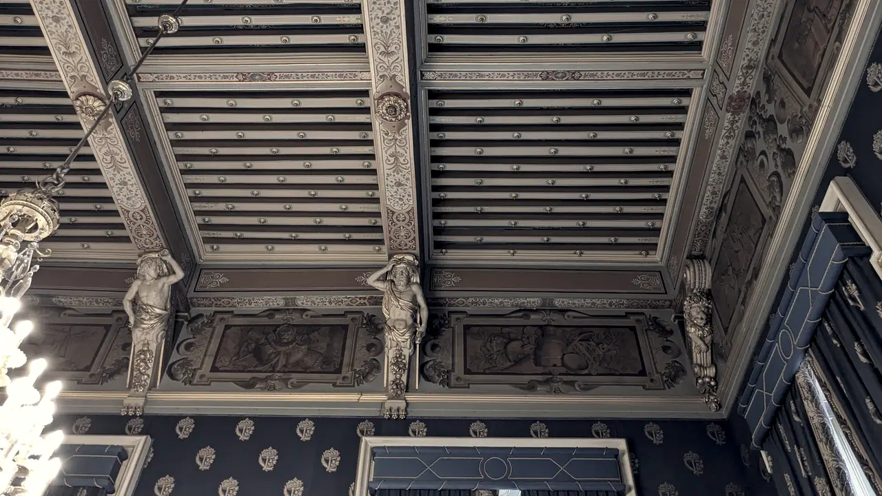 Plafond à la française d'un salon ancien : poutres et solives ornées de rinceaux, portées par des atlantes sculptés, avec un lustre sur la gauche.