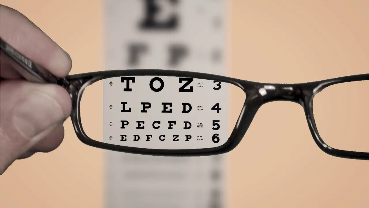 Main tenant une paire de lunettes devant un tableau de lecture flou : derrière les verres, les lignes de lettres redeviennent nettes jusqu'aux plus petites.