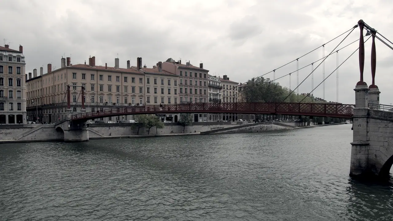 Passerelle suspendue franchissant une rivière, avec l'alignement des façades anciennes le long du quai au fond