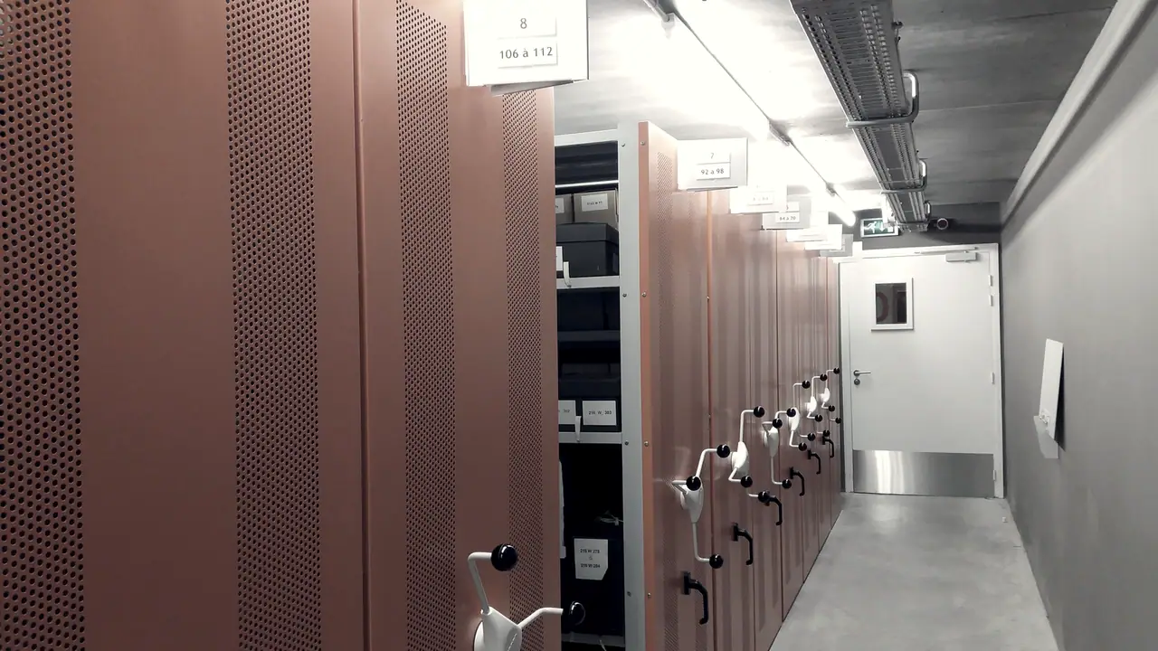 Rayonnages mobiles perforés alignés dans un couloir d'archives, avec leurs volants de manoeuvre et des étiquettes numérotées