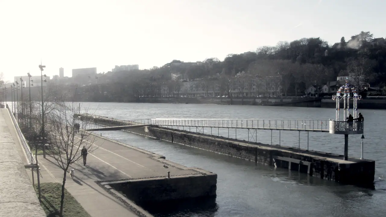 Môle de pierre et sa passerelle métallique avançant dans la rivière, avec des coteaux boisés au fond
