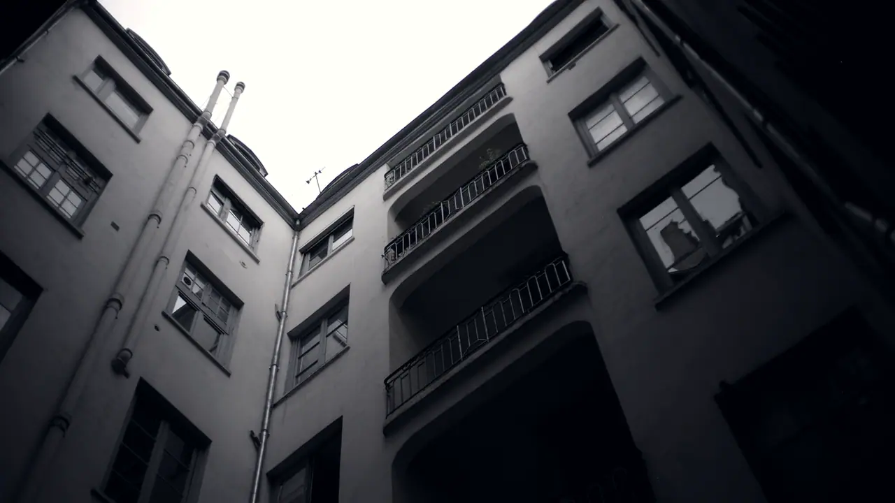 Cour intérieure étroite vue en contre-plongée, entourée de façades percées de fenêtres, avec une colonne de balcons à garde-corps de fer et un pan de ciel au-dessus des toits.