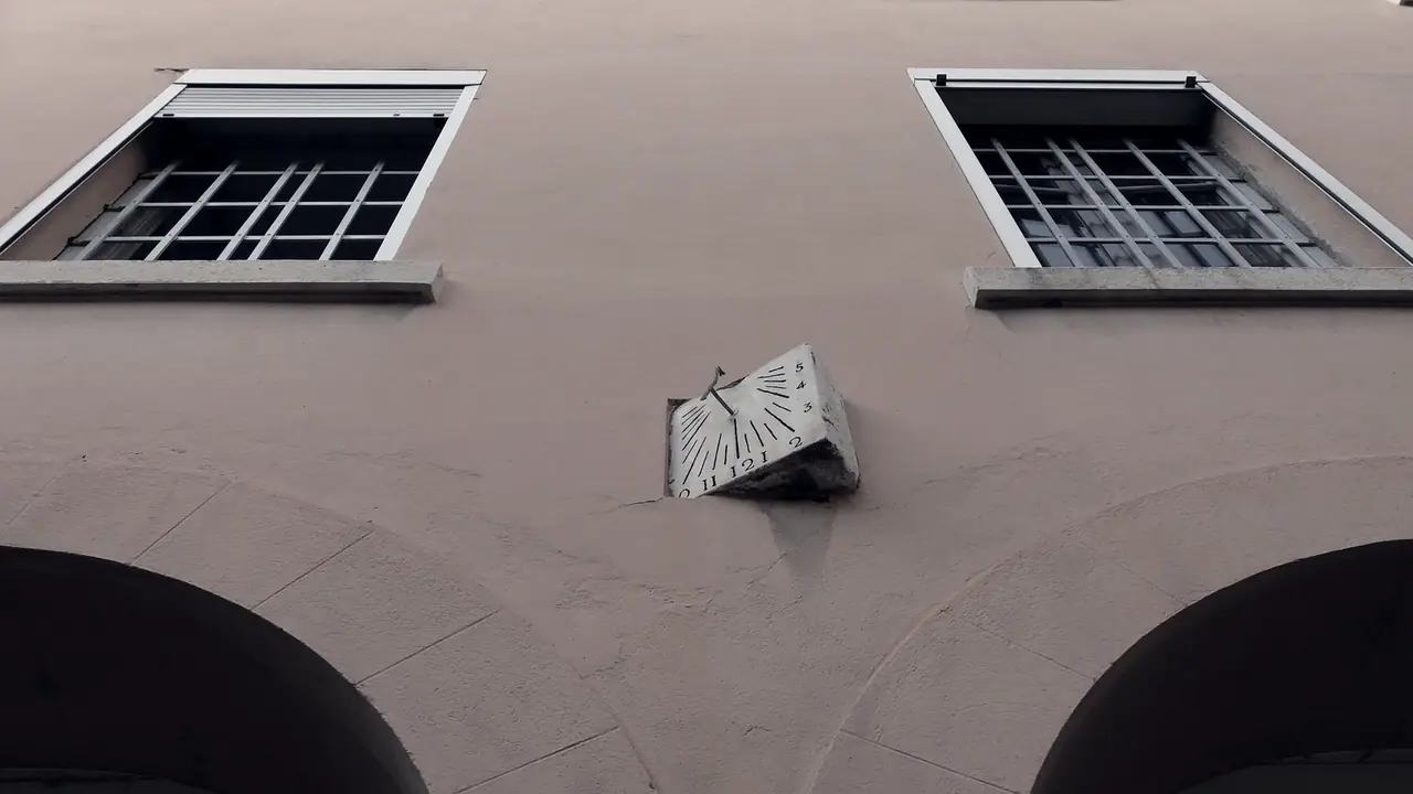 Cadran solaire fixé sur le mur d'un bâtiment, sous deux fenêtres à barreaux et au-dessus de deux arcades, avec ses heures gravées autour du style.