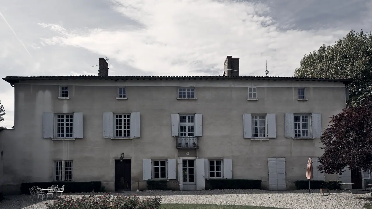 Grande maison bourgeoise à deux étages, ses volets de bois ouverts, devant une cour de gravier bordée de pelouse et d'un arbre.