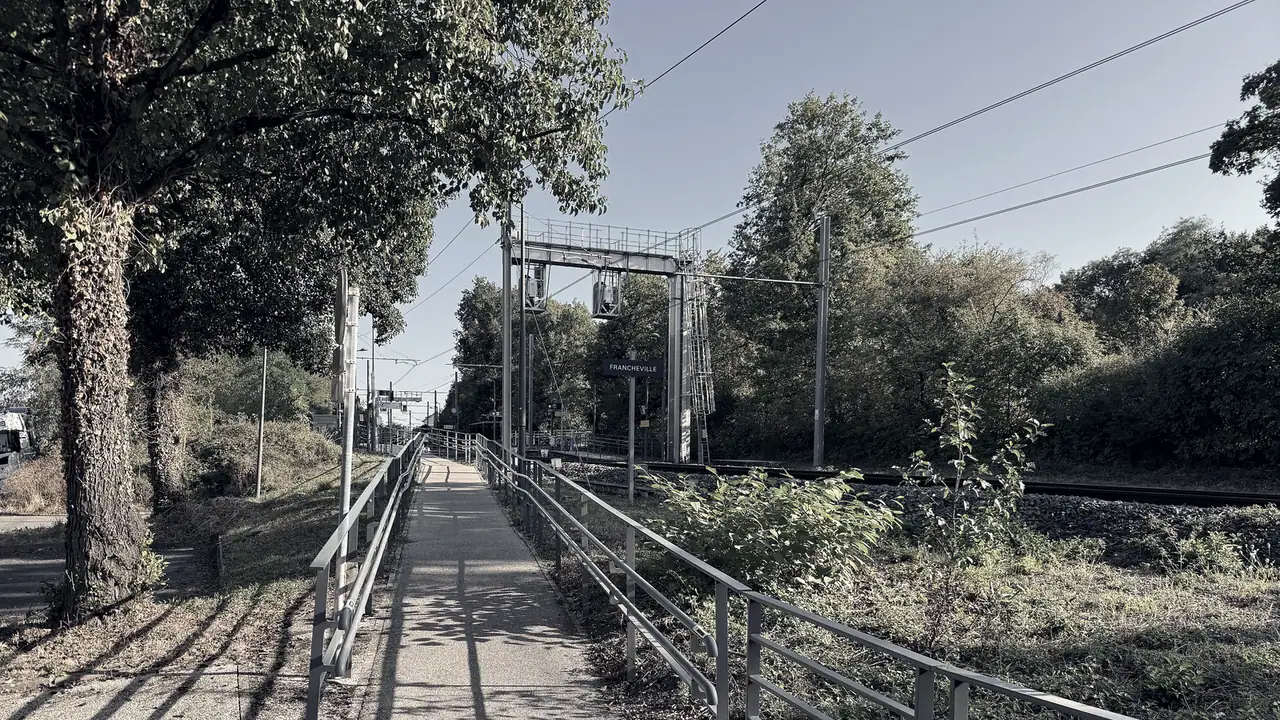 Chemin bitumé bordé de rambardes métalliques longeant une voie ferrée, sous les câbles caténaires et les arbres en plein été.