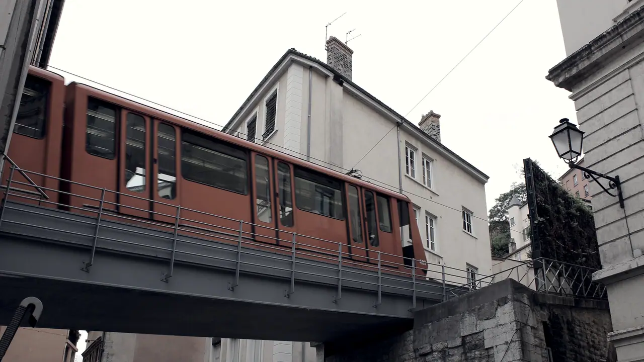 Rame du funiculaire de Saint-Just passant sur un pont métallique au-dessus d'une rue, devant un immeuble ancien et une lanterne fixée à l'angle d'un mur.