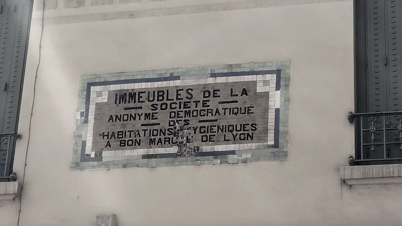 Plaque de mosaïque encadrée, scellée sur un mur crépi entre deux fenêtres à garde-corps, annonçant des immeubles d'habitations à bon marché.