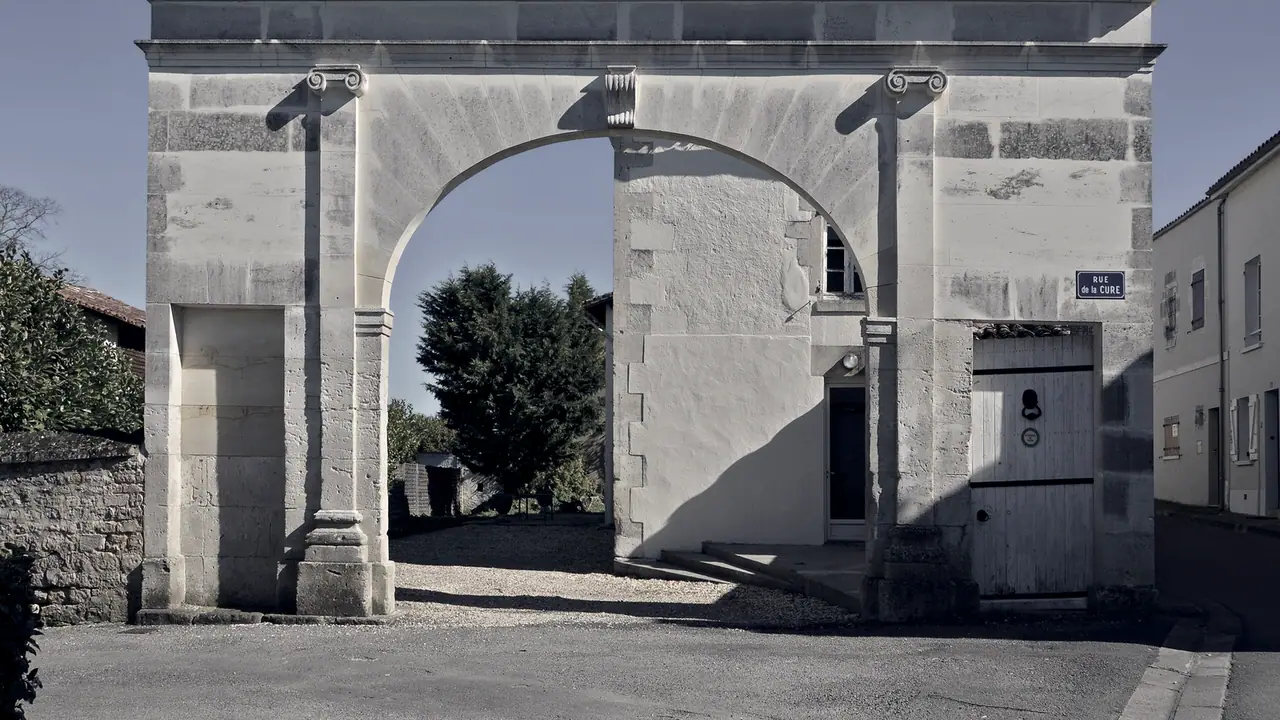 Portail ancien en pierre de taille : la grande arche reste ouverte sur une cour plantée, tandis que la baie de gauche a été murée de pierre.