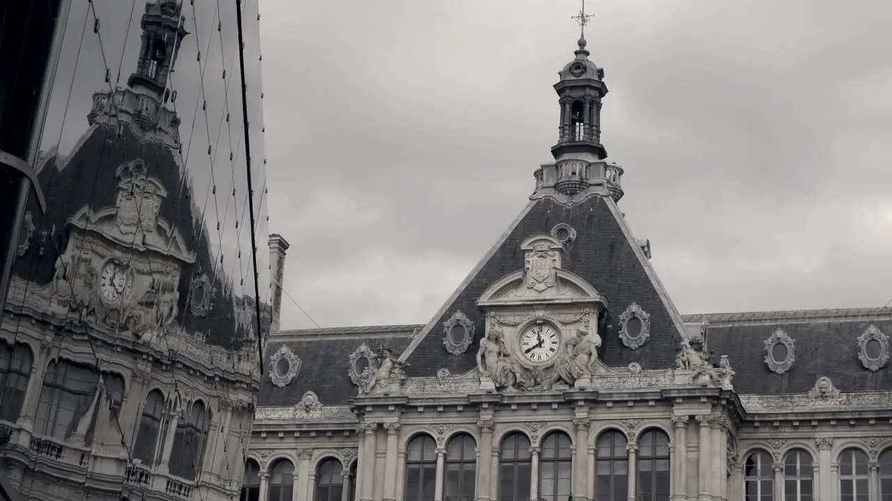 Toiture sculptée d'un palais ancien avec son horloge et son clocheton, reflétée dans la façade de verre voisine sous un ciel couvert.