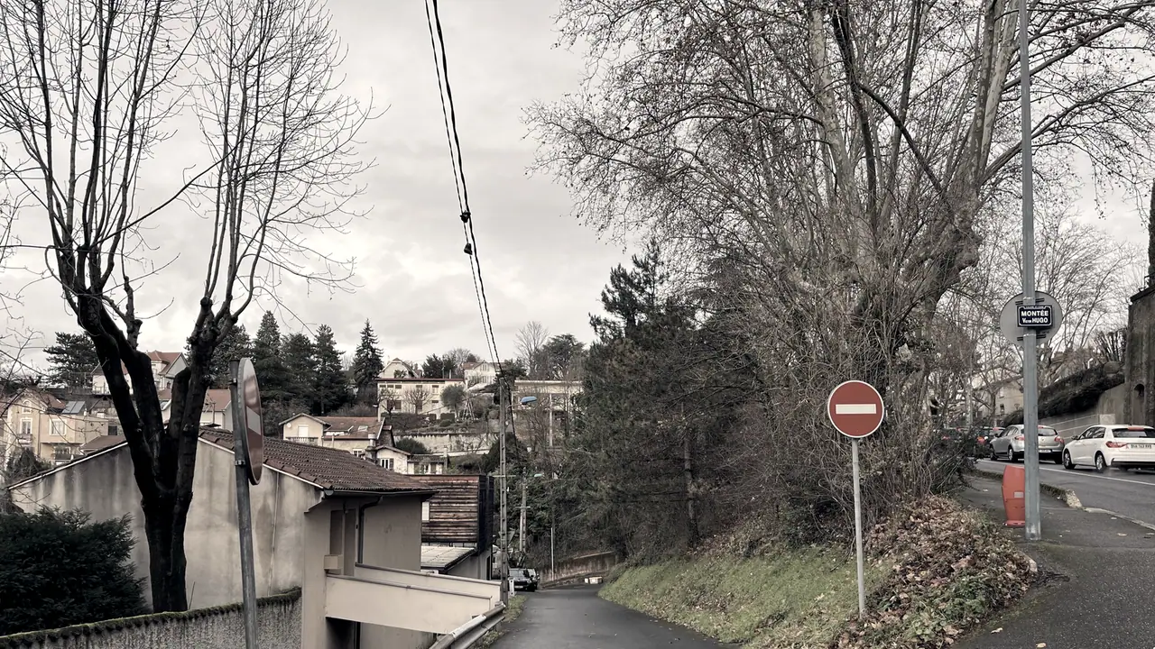 Deux voies qui divergent au sommet d'une montée, un panneau de sens interdit et un miroir de carrefour, maisons perchées au fond.