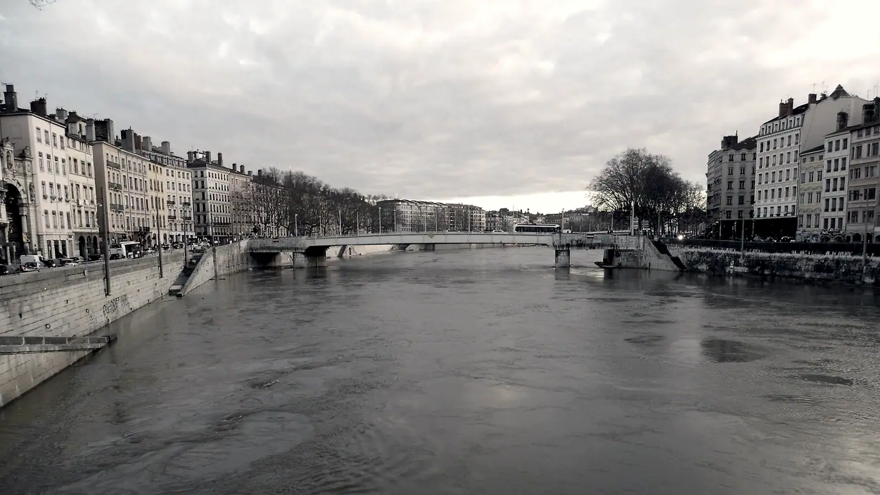 Rivière large et trouble en crue entre deux quais de pierre, un pont bas barrant l'horizon devant les façades alignées.