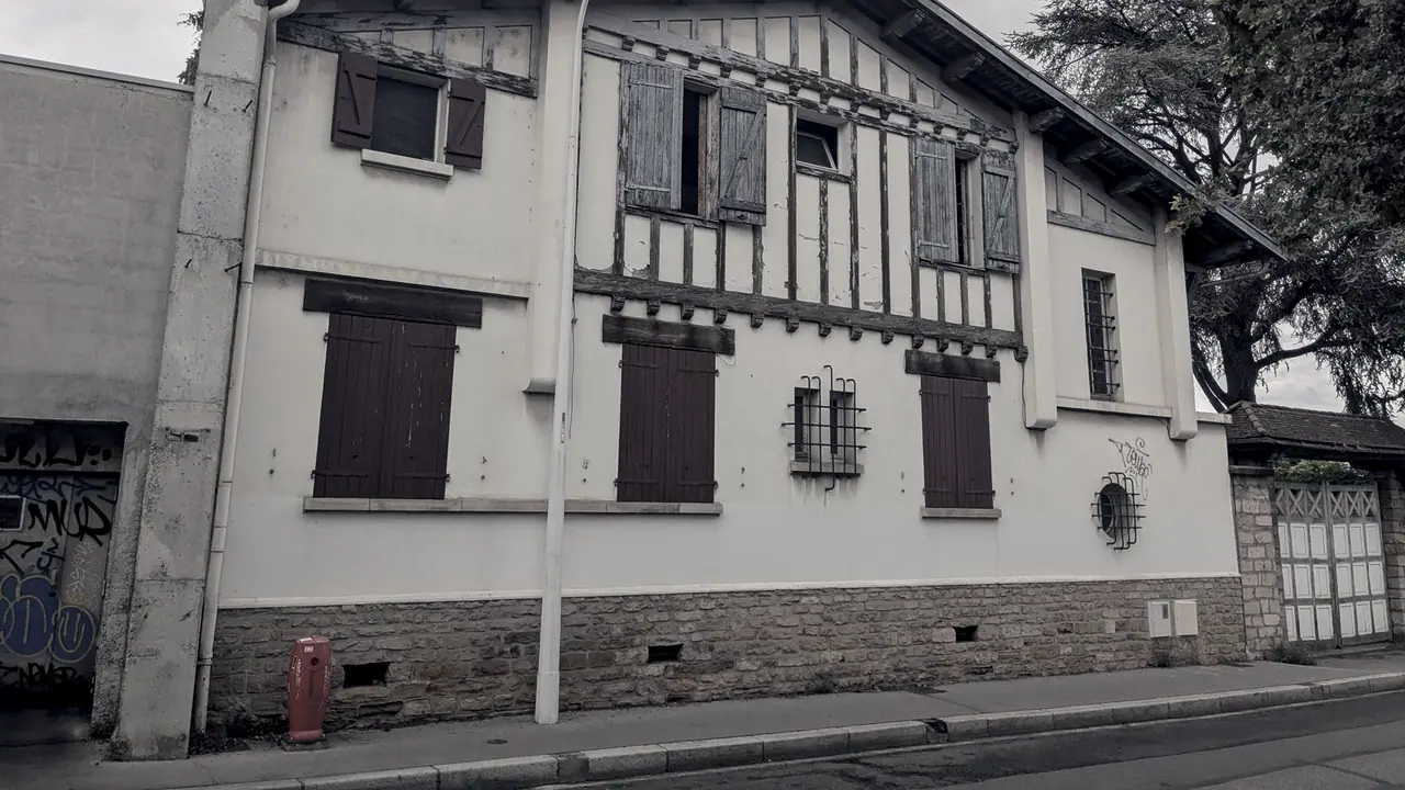 Maison à deux niveaux vue depuis le trottoir, avec des volets de bois, un pan de colombages à l'étage et un soubassement en pierre.