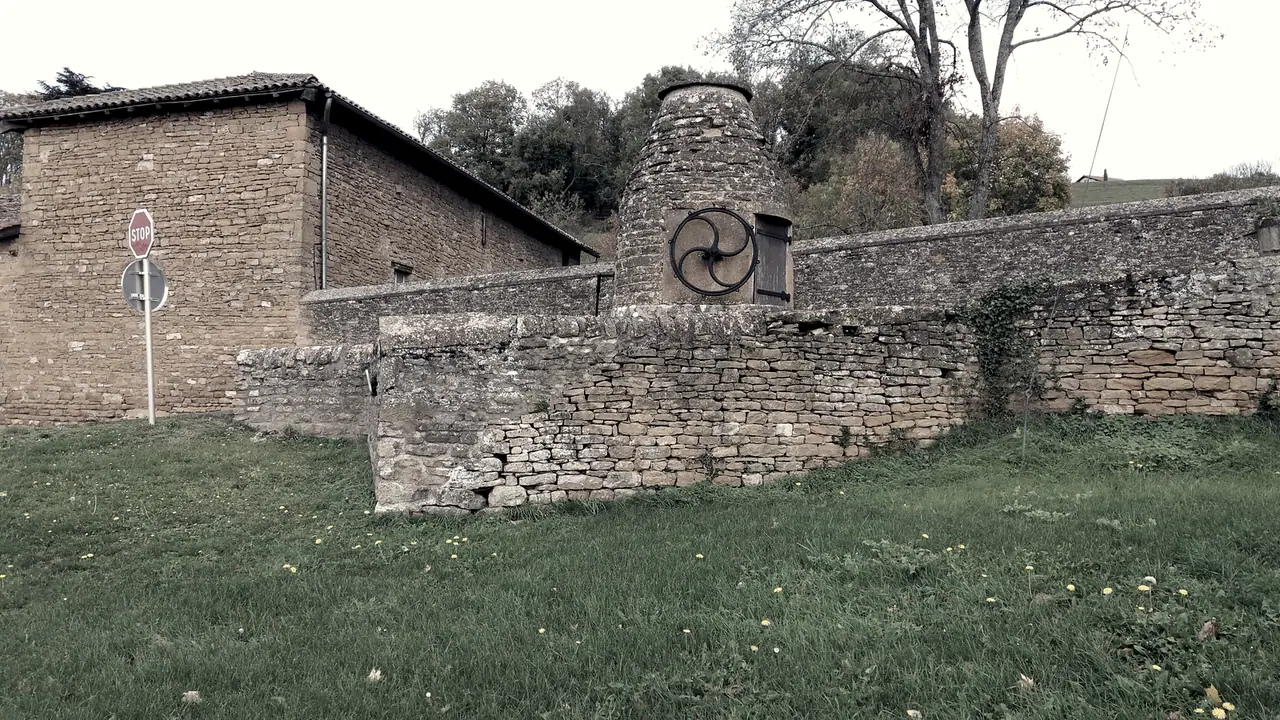 Puits couvert en pierre dorée, coiffé d'un cône de pierres et muni de sa roue de fer, pris dans un mur de clôture près d'une maison, au bord d'un pré.